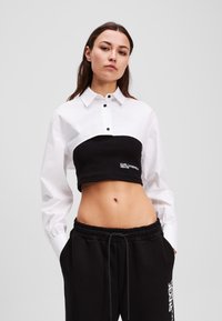 White button-up shirt with a black cropped ribbed bodice featuring logo. Black sweatpants with an elastic waistband and side pockets.
