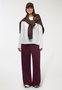 Biała bluzka z długim rękawem, brązowy sweter przewiązany na ramionach, bordowe spodnie szerokie w nogawkach i brązowa czapka, w komplecie z szarymi sneakersami.