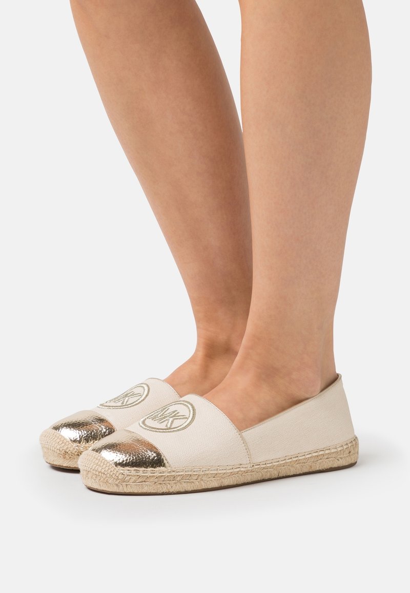 Pieds portant des espadrilles beiges avec embouts métalliques dorés et logo circulaire brodé dessus, posés sur un fond clair uni.