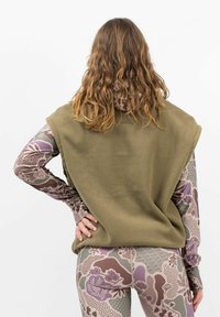 Olivgrüner Fleece-Westen mit weitem Armloch, kombiniert mit einem langärmeligen gemusterten Hemd in verschiedenen Lila- und Beigetönen, vor einem weißen Hintergrund.