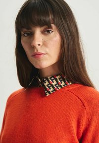 Pull en maille orange avec un col à motif présentant des designs géométriques en beige, rouge et noir. Texture lisse et coupe décontractée.