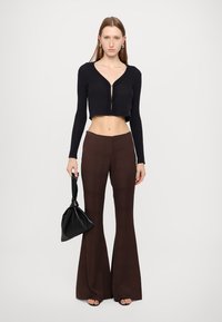 Black ribbed cropped cardigan and brown flared pants with a grid pattern. Model holds a black handbag, with open-toe sandals.