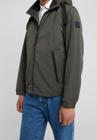 Olive green zip-up jacket with hood, featuring black zippered pockets and straps on shoulders, made of lightweight fabric.
