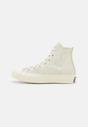 Kremowy sneaker za kostkę z subtelnym falistym wzorem linii, białe sznurowadła, gumowa podeszwa i czarne logo na pięcie, widoczny z profilu bocznego.
