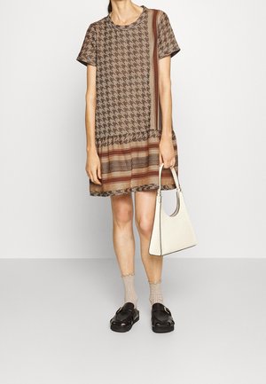 Kvinde iført en brun mønstret kortærmet kjole, som holder en hvid håndtaske, kombineret med beige sokker og sorte slip-on sko.