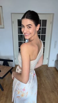 Νεαρή γυναίκα που χαμογελά κοιτάζοντας πίσω από τον ώμο της, φοράει ένα μπεζ τοπ με έναν ώμο και μια λευκή φλοράλ φούστα, στέκεται σε δωμάτιο με ξύλινο πάτωμα.