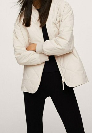 Nainen pukeutuneena kellertävänvalkoiseen tikattuun takkiin mustan topin ja housujen päällä, seisoo käsivarret ristissä yksivärisen taustan edessä.