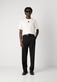 Weißes Kurzarmhemd mit kleinem schwarzem Logo, kombiniert mit schwarzen Hosen und schwarzen Schuhen. Model trägt dunkle Sonnenbrillen.