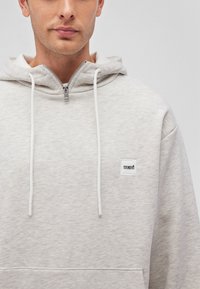 Light gray zip-up hoodie made of soft material, featuring a white logo patch, drawstrings, and a kangaroo pocket.