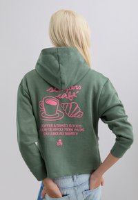 Sudadera verde con diseño gráfico rosa de una taza de café y un croissant, con texto. Material de algodón, diseño corto, capucha ajustable.