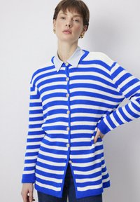 Cardigan rayé bleu et blanc avec une fermeture à bouton, texture côtelée et un col bleu clair. Des boutons dorés remarquables ornent l'avant.