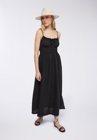 Abito maxi nero con spalline regolabili, corsetto arricciato e scollatura volants. Indossato con sandali neri con cinturini e un cappello di paglia bianco.