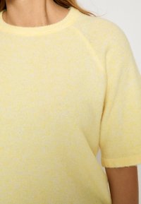 Dzeltena adīta džemperis ar apaļu kaklu, īsām raglāna piedurknēm un mīkstu tekstūru ar subtilu plankumainu rakstu.