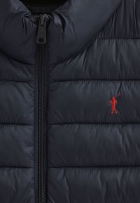 Granatowa ocieplana kamizelka z wysokim kołnierzem i pionowym przeszyciem. Zamek błyskawiczny na środku oraz czerwony haftowany logo golfisty na lewej piersi.