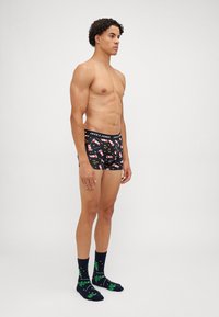 Schwarze Boxershorts mit bunten Mustern von Santa-Mützen und Geschenken; getragen mit blauen Socken, die grüne Cartoon-Bäume und Textakzente zeigen.