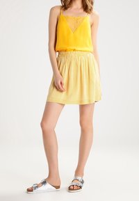 Camisole jaune avec bordure en dentelle associée à une jupe plissée à rayures jaunes. Sandales argentées complètent le look. Design simple et décontracté.