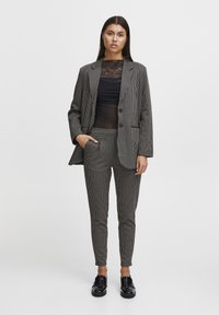 Melnbalts houndstooth uzvalks ar pielāgotu žaketi un sašaurinātiem biksēm, zem kura ir caurspīdīga melna blūze, valkāts ar spīdīgām melnām kurpēm.