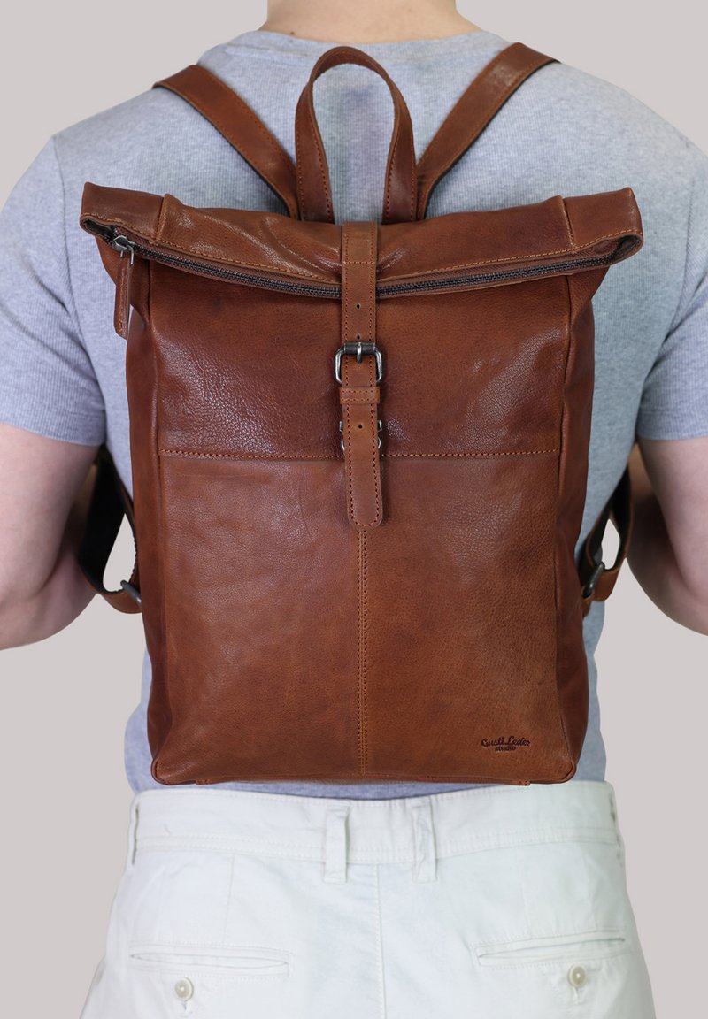Zaino in pelle marrone con design a roll-top, hardware in metallo e spallacci regolabili. Presenta una tasca frontale e una texture liscia.