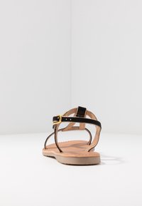 Black strappy sandal with a gold buckle, flat sole, and a brown insole. The straps are made from soft material with a minimalistic design.