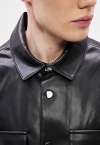 Melna ādas jaka ar pogājamu apkakli, sudraba detaļām un gludu tekstūru. Ir priekšējās kabatas detaļa.