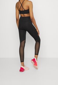 Mallas deportivas negras con paneles de malla y una cinturilla alta, combinadas con zapatillas rosas y beige que presentan suelas texturizadas.