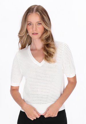 Ung kvinde med bølget blondt hår iført en hvid striktop med korte ærmer og en V-hals med et perforeret mønster.