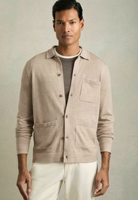 Cárdigan de punto beige con cuello abierto, botón frontal y dos bolsillos delanteros, usado sobre una camisa gris, combinado con pantalones claros.