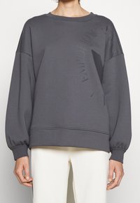 Sudadera gris de gran tamaño con mangas largas y abullonadas. Presenta un print texturizado del logo en la parte frontal y puños y dobladillo de canalé.
