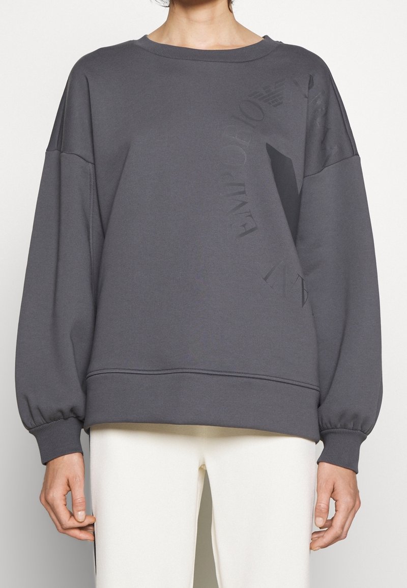 Sudadera gris de gran tamaño con mangas largas y abullonadas. Presenta un print texturizado del logo en la parte frontal y puños y dobladillo de canalé.