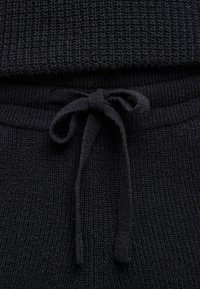 Czarny sweter z dzianiny o prążkowanej fakturze, z zapięciem na sznurek. Materiał jest miękki, o jednolitym wzorze na całej powierzchni.