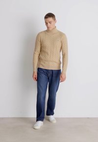 Suéter beige de punto trenzado con cuello alto, combinado con jeans azul oscuro y zapatillas blancas. Fondo liso, modelo de pie con las manos a los lados.