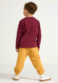 Burgundy pitkähihaisessa paidassa, keltaisissa verkkareissa ja valkoisissa lenkkareissa. Pehmeä kangas, rento malli, joustavat hihansuut housuissa, yksinkertainen muotoilu, ilman kuvioita.