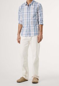 Muž v světle modré a bílé kostkované košili, bílých kalhotách a béžových slip-on botách, stojící proti jednobarevnému pozadí.