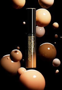 Il correttore Luminous Lift 24HR MAC Studio Radiance in un tubo trasparente con un tappo nero, circondato da sfere lucide beige e marroni su uno sfondo nero.