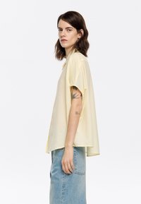 Loose-fit, short-sleeve shirt in light yellow textured fabric with a subtle check pattern, featuring a collar and relaxed silhouette.