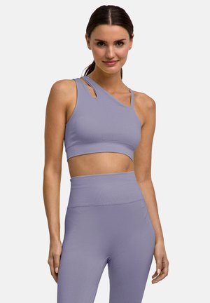 Vrouw met donker haar draagt een lichtpaarse asymmetrische sportbeha en hoge taille leggings, staat met één ontspannen hand langs haar zij.