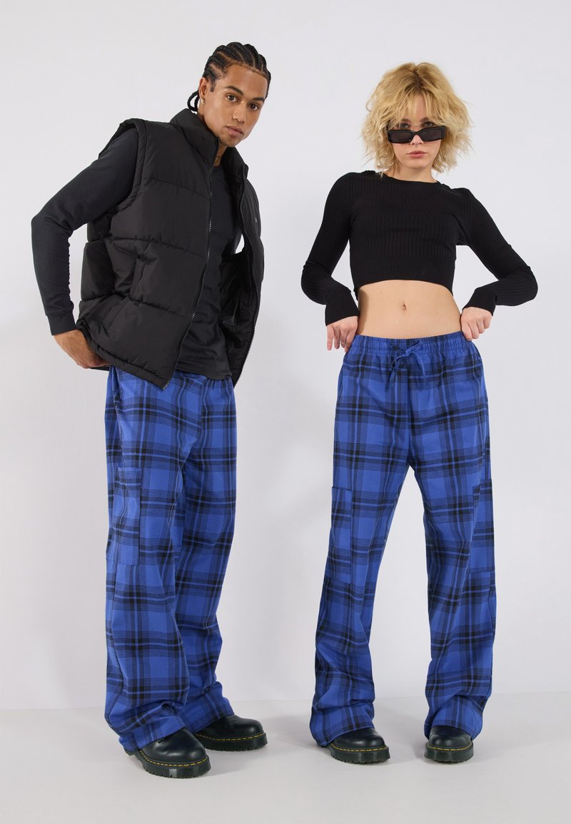 Zwarte puffervest, lange mouwen zwart top, baggy blauwe ruitbroek, zwarte schoenen met gele stiksels; twee modellen staan tegen een witte achtergrond.