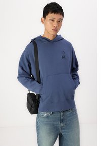 Μπλε βαμβακερή κουκούλα με μπροστινή τσέπη και μικρό γραφικό λογότυπο. Φοριέται με ανοιχτόχρωμες μπλε τζιν και μαύρη τσάντα χιαστί.