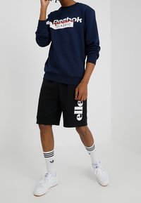 Marinblå sweatshirt med "Reebok Classic"-logotyp, ihop med svarta shorts med vit text. Vita sneakers och randiga strumpor fulländar looken.