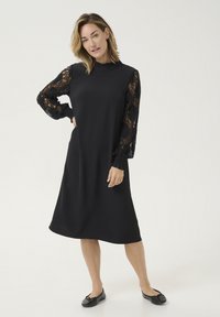 Abito nero con maniche lunghe in pizzo e polsini arricciati, scollo ruffles e taglio morbido, abbinato a scarpe basse nere.