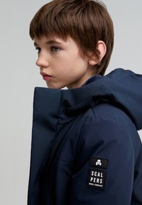 Mørkeblå hættetrøje med høj krave, lavet af glat materiale. Udstyret med et sort mærkat med et kranie logo på ærmet.