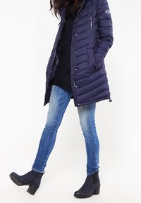 Mörkblå vadderad jacka med huva, med horisontella sömmar, dragkedjeförsedda fickor och ett märkesmärke. Bärs över en svart tröja och blå jeans.