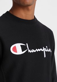 Felpa nera con collo a girocollo a costine; presenta un grande logo "Champion" ricamato in bianco e un'emblema rosso e bianco. Tessuto morbido.
