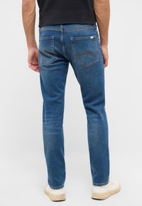 Męskie niebieskie dżinsy o prostym kroju, z tylnymi kieszeniami, pomarańczowymi szwami i lekko podniszczoną fakturą. Noszone z białymi trampkami.