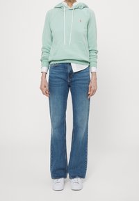 Mint green hoodie with a kangaroo pocket, white drawstrings, layered over a white shirt. Paired with flared blue jeans and white sneakers.