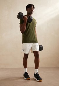 Grön rutig idrottströja, vita shorts med logotyp, håller två svarta hexagonala hantlar, bär svarta sneakers med vita detaljer.