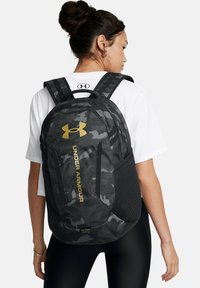 Sac à dos camouflage noir avec un logo Under Armour doré, poches latérales en mesh, bretelles rembourrées et un design élégant et ergonomique.