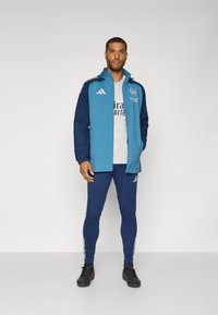 Blauwe en marineblauwe sportjack met capuchon, gecombineerd met bijpassende strakke broek. Bevat een logo op de borst en strepen op de mouwen en broek. Zwarte schoenen.