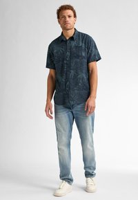 Camisa de botones de manga corta en azul oscuro con un patrón de hojas, combinada con jeans de azul claro y deportivas blancas. Detalle de tejido texturizado visible.