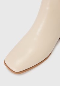 Botte courte en simili cuir beige avec un bout carré, texture lisse et un talon bas empilé. La semelle présente un accent marron clair.